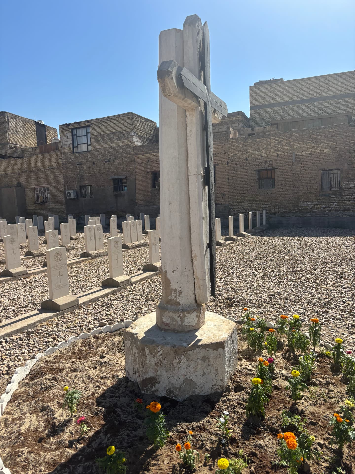 The Cross of Sacrifice at close quarters, the upper section bolted to the lower and propped by a painted metal cross, standing on a rough concrete base.