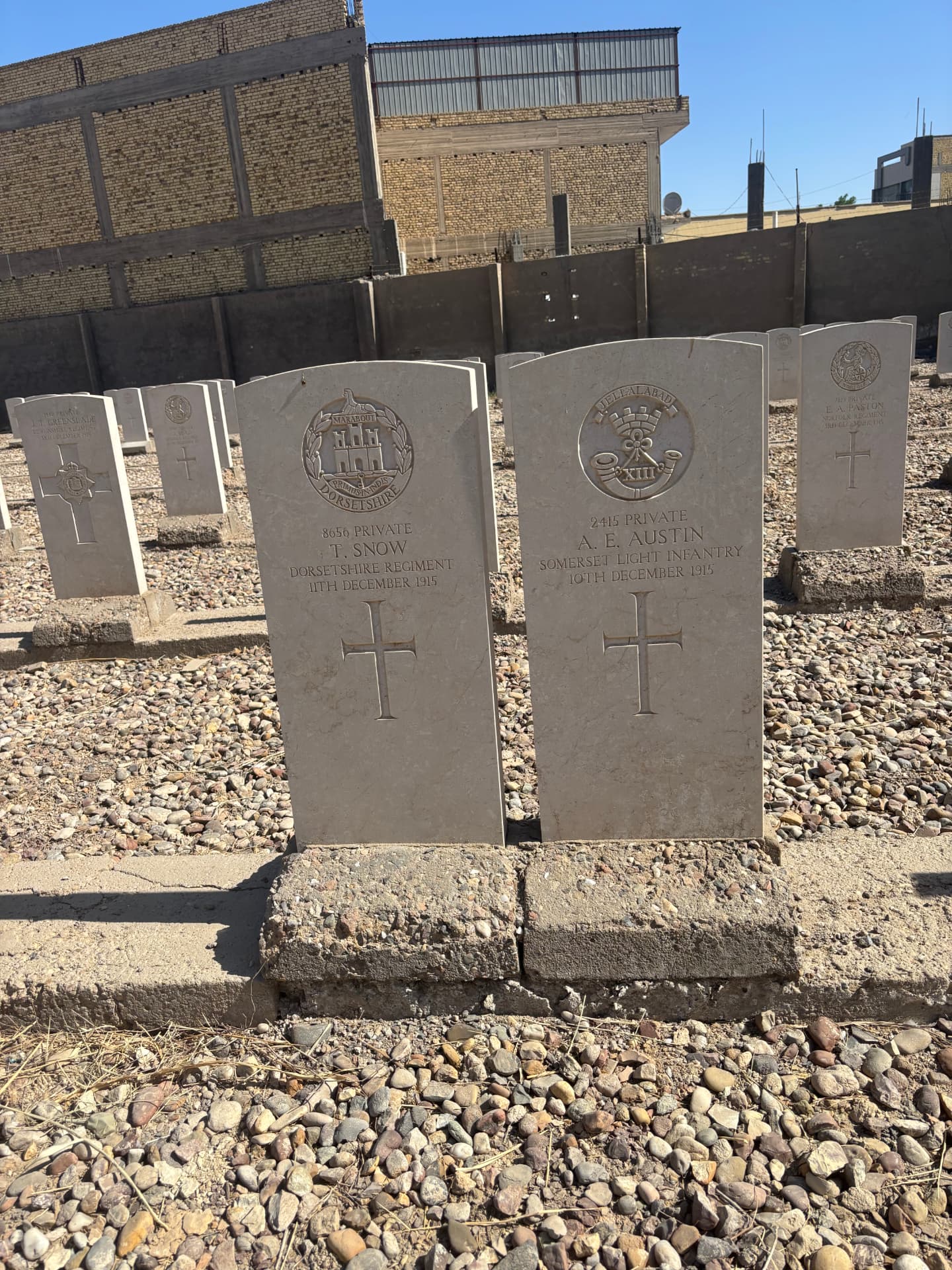 The graves of 8656 Private T. Snow, Dorsetshire Regiment, died 11 December 1915, and 2415 Private A. E. Austin, Somerset Light Infantry, died 10 December 1915, with the town pressing up against the cemetery wall behind.