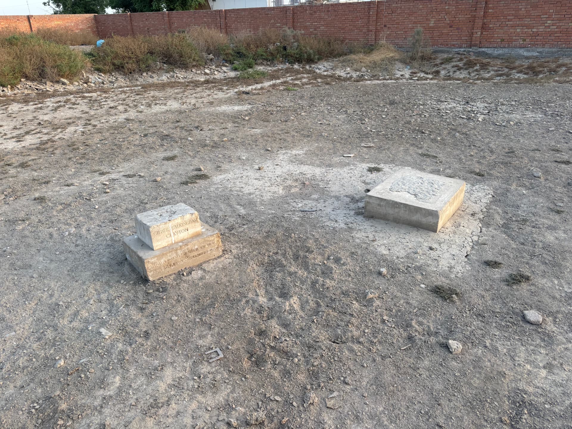 The Clayton grave in its wider setting at Ma'asker al Raschid, the inscribed stone at left and a stripped adjacent grave slab at right, with the modern brick perimeter wall beyond.
