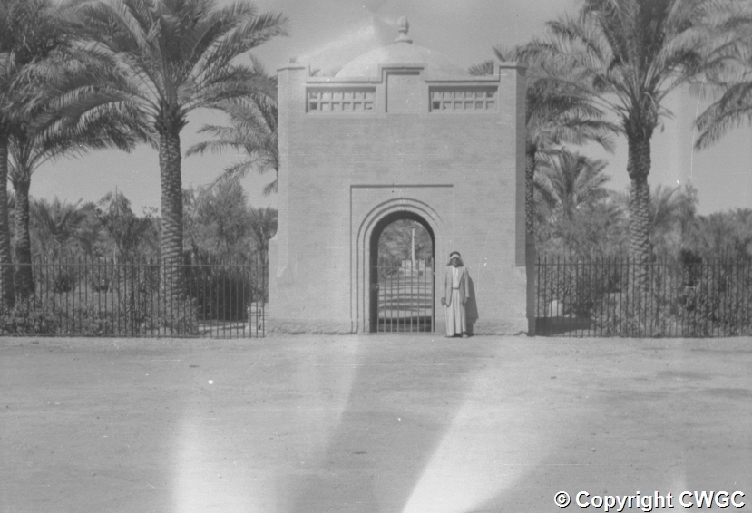 The gatehouse photographed from the approach road, with a local figure standing beneath the archway and the Cross of Sacrifice visible through the gate beyond. © CWGC.