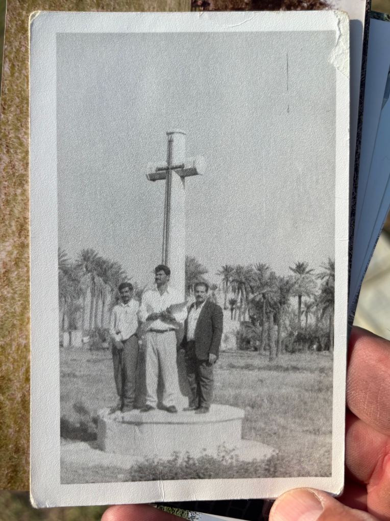 A photograph from the Hasan family archive, three men gathered at the base of the Cross, the bronze sword still mounted on the shaft and date palms visible behind. Taken after the original headstones had been removed.