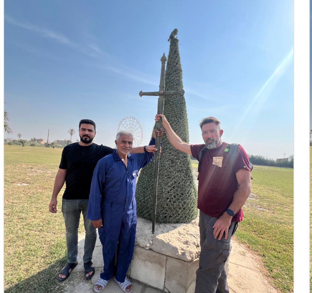 The site of the Cross of Sacrifice today, the pedestal alone remaining on a wide expanse of cleared ground. Mr. Hasan has built up and dressed the pedestal with greenery in the form of the original Cross. A modern road and street lighting are visible along the boundary beyond.