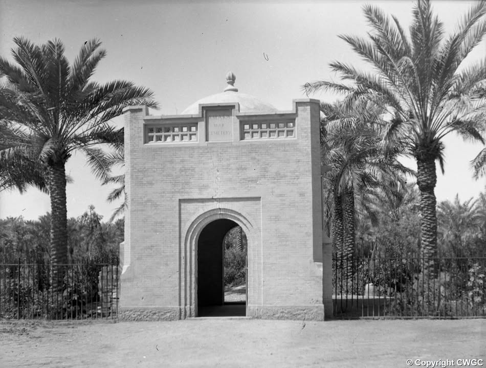 The original gatehouse, with its inscribed lintel and domed cupola, framed by date palms and the iron perimeter railings of the cemetery. © CWGC.