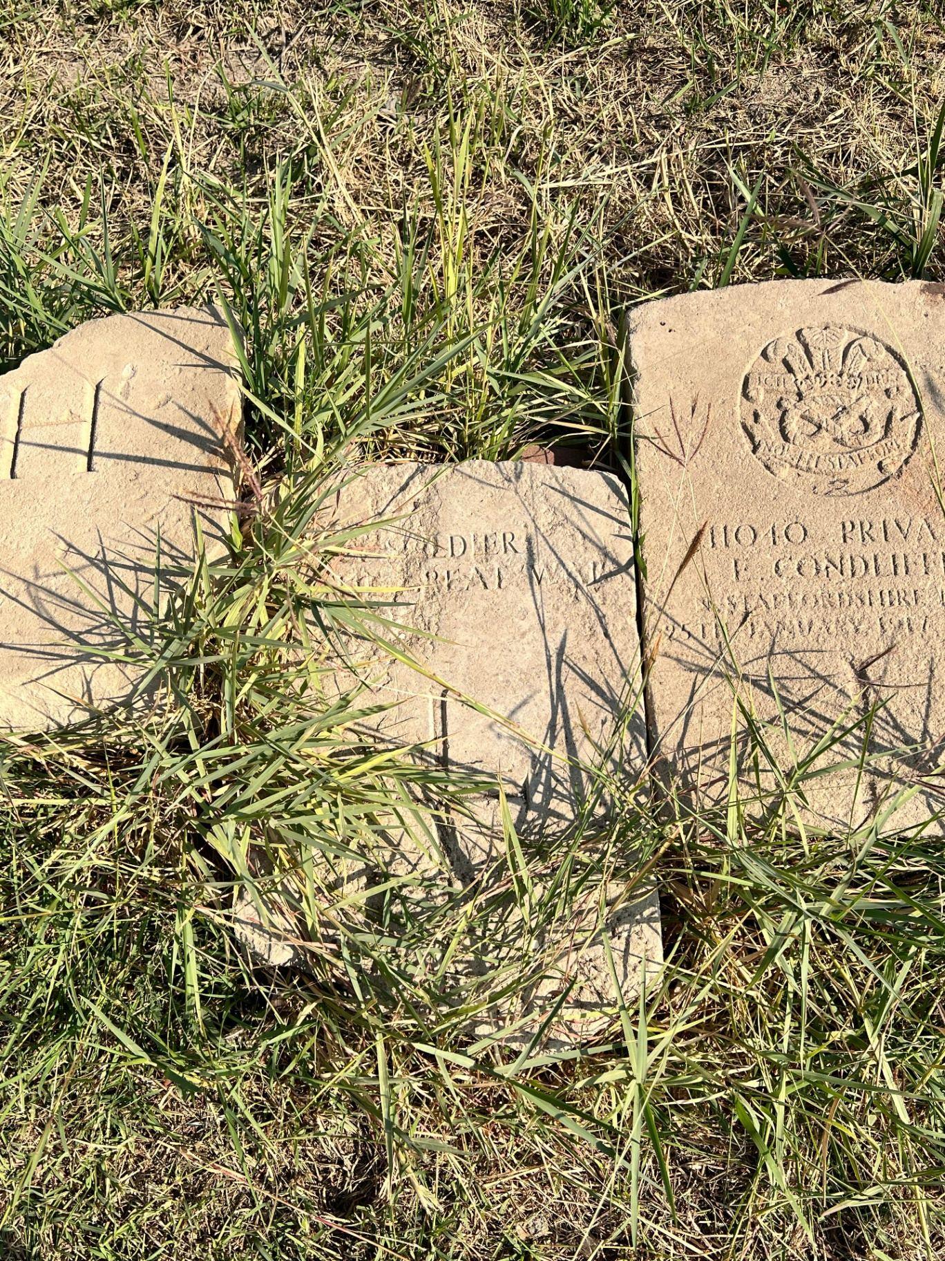 Close-up of surviving headstone fragments in the grass, including the regimental badge of the North Staffordshire Regiment on the marker for 11040 Private E. Condliffe of the North Staffordshire Regiment, killed 25 January 1917.