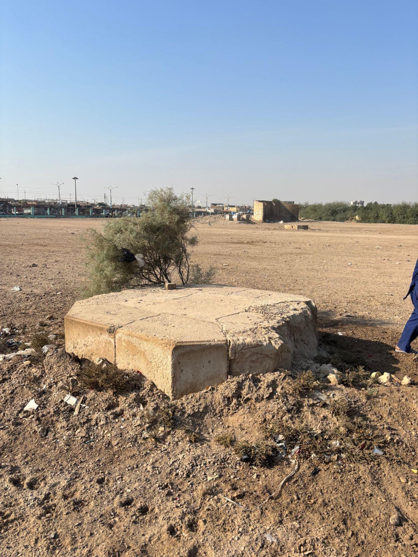 The surviving pedestal of the Indian Forces Memorial sitting alone on the bare ground of the former cemetery, with the surrounding city visible in the distance