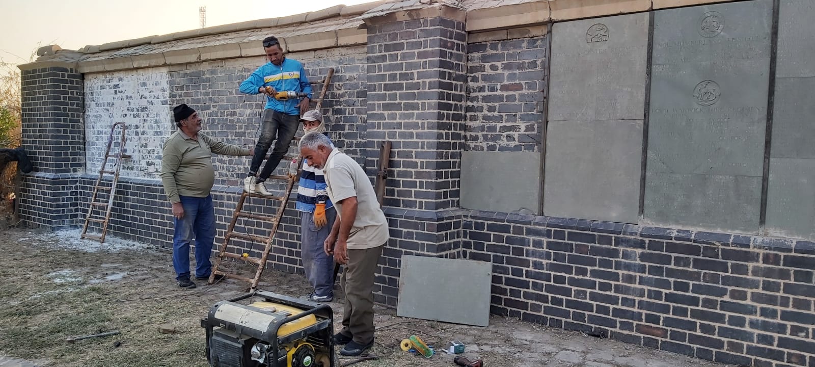 Mr Hasan and members of his team installing panels on the renovated Memorial Wall, working from a wooden ladder with regimental crests visible at right.