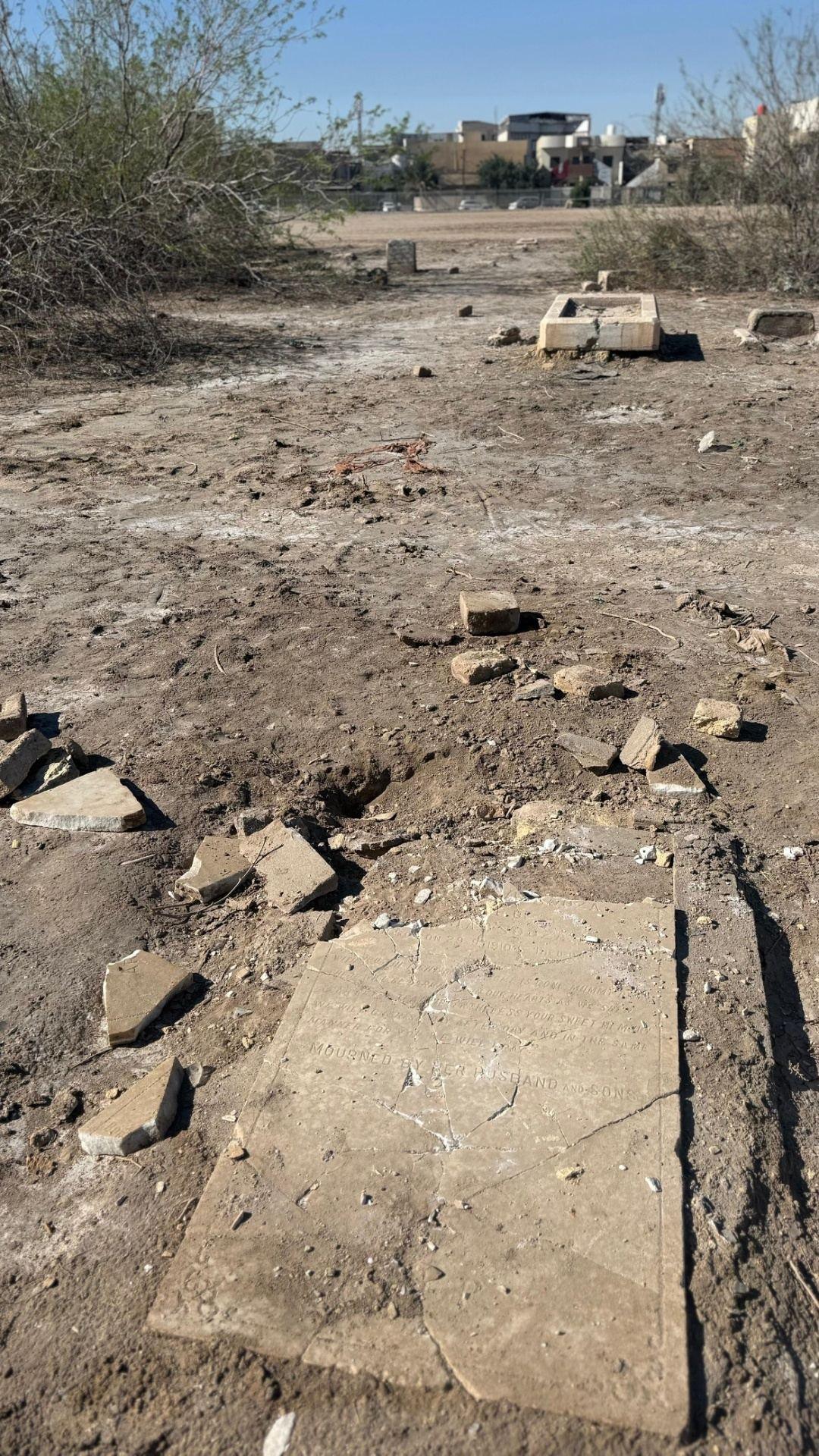 A broken and cracked civilian grave slab lying flat on the dusty ground in Basra War Cemetery, surrounded by scattered fragments of stone, with the inscription "Mourned by her husband and sons" still partially legible.