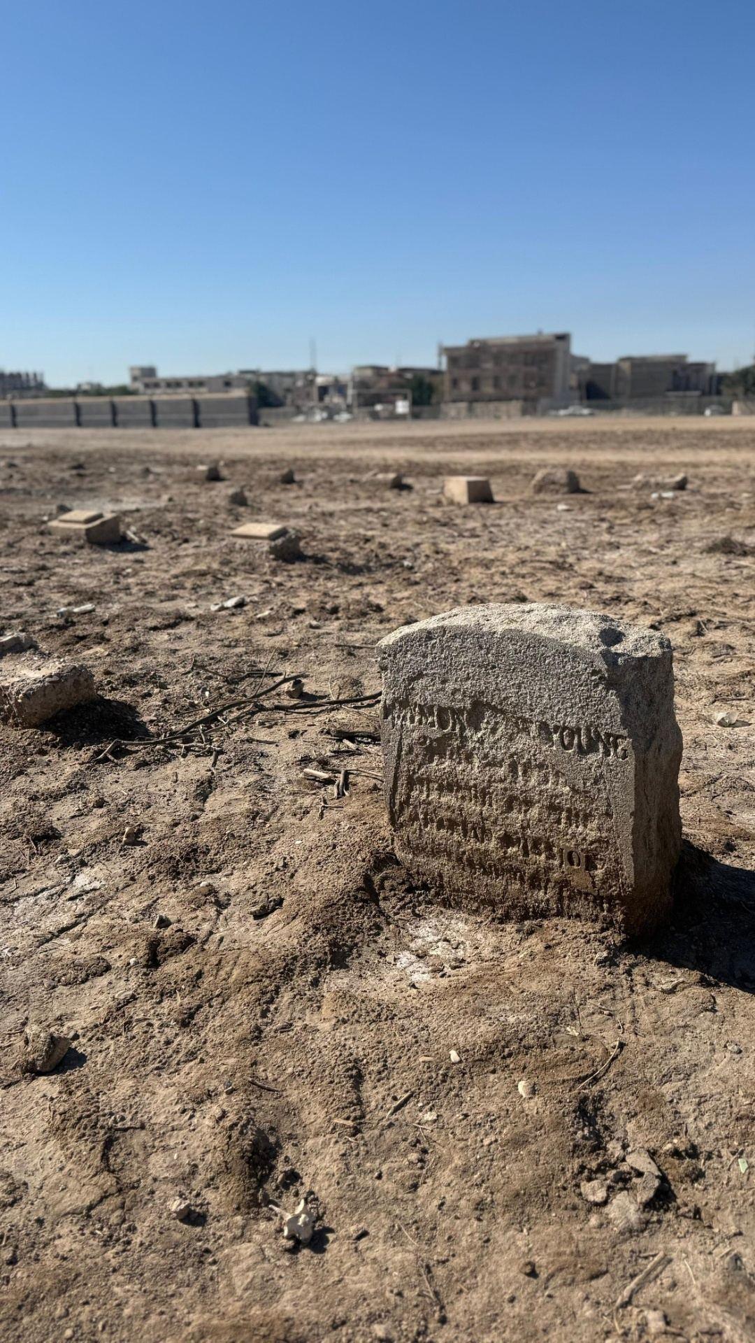 A weathered civilian headstone bearing the name "Young" leaning in the dry earth of the civilian cemetery at Basra, with other broken grave markers and the modern Basra skyline behind a perimeter wall in the background.