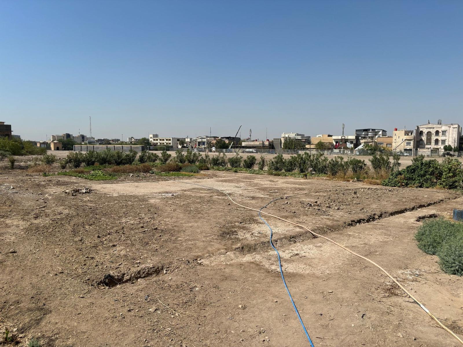 The site of the RAF Peace cemetery at Basra War Cemetery, now a bare patch of cracked earth with hosepipes running across it, fringed by low vegetation, with the modern Basra skyline visible in the background.