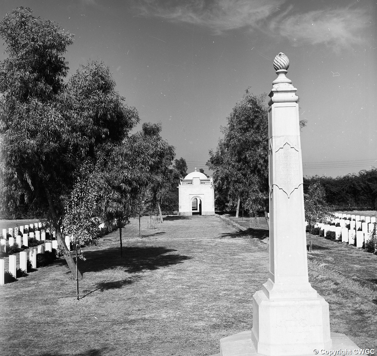 View along the central avenue of the Indian Forces side of Basra War Cemetery, with the obelisk in the foreground and the domed gatehouse in the distance, framed by mature trees and rows of headstones. © CWGC.
