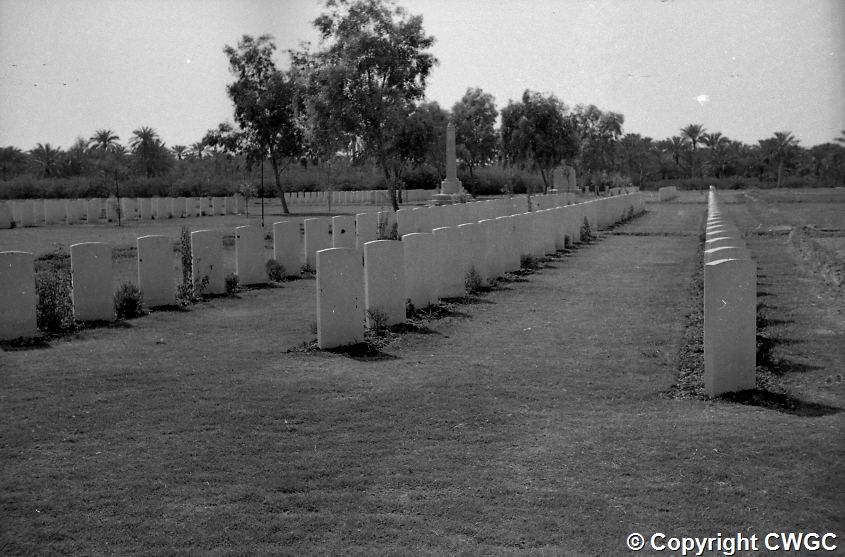 Long rows of headstones at the Indian Forces side of Basra War Cemetery, with the obelisk visible in the distance among palms and trees. © CWGC.