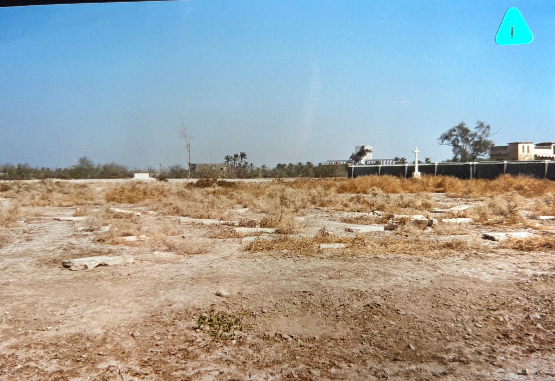 Basra War Cemetery in 1997, the grounds reduced to bare earth and scrub, with broken stone fragments scattered across the foreground and the Cross of Sacrifice still standing in the distance against a perimeter screen. © CWGC.