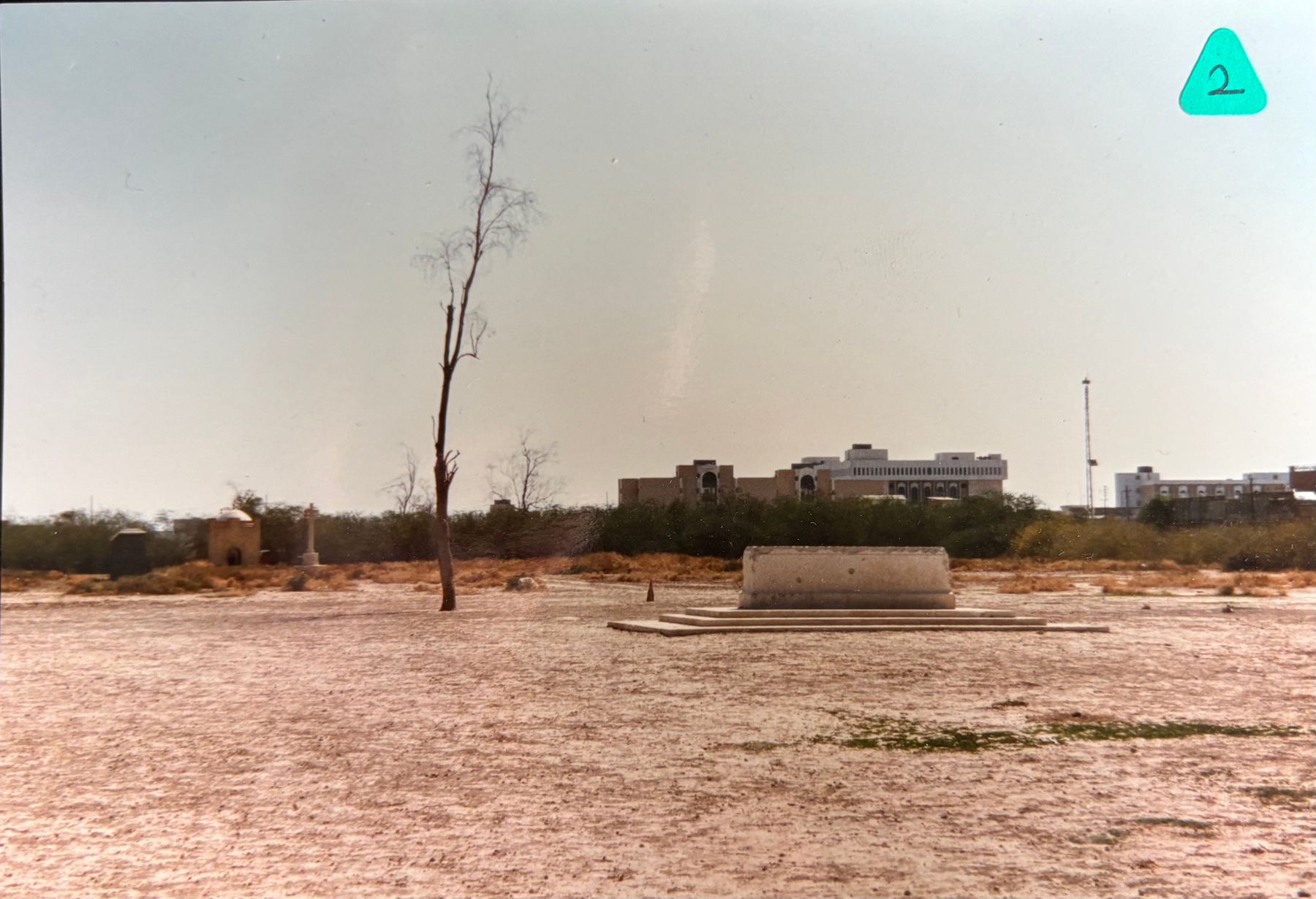 Basra War Cemetery in 1997, the Stone of Remembrance standing on its tiered platform on bare ground, with the Cross of Sacrifice and a surviving brick gatehouse visible in the distance and the modern city rising beyond. © CWGC.