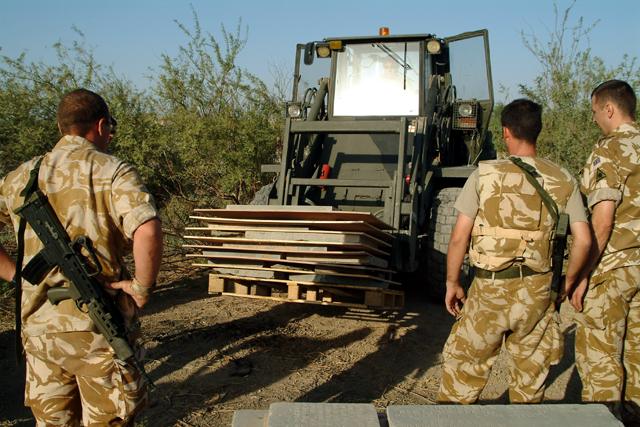 A military forklift moves a stack of memorial panels strapped to a pallet through the cemetery in 2003, with armed soldiers providing close protection during the recovery operation.