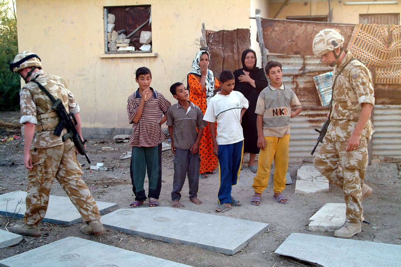 Local Iraqi families watch as British soldiers move across the cemetery in 2003, with displaced memorial panels and a fragmentary headstone visible in the foreground beside their home built up against the cemetery boundary.