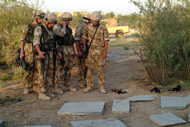 A patrol of British soldiers pause beside fallen and broken memorial panels lying on the bare ground in 2003, taking in the scale of the damage at the cemetery before the salvage operation begins.
