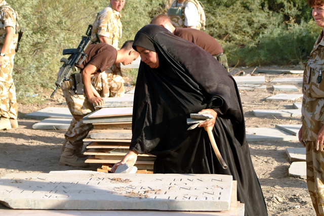 An Iraqi woman gently brushes sand from a recovered headstone laid on the ground in 2003, with British soldiers stacking further panels on pallets behind her during the salvage operation.