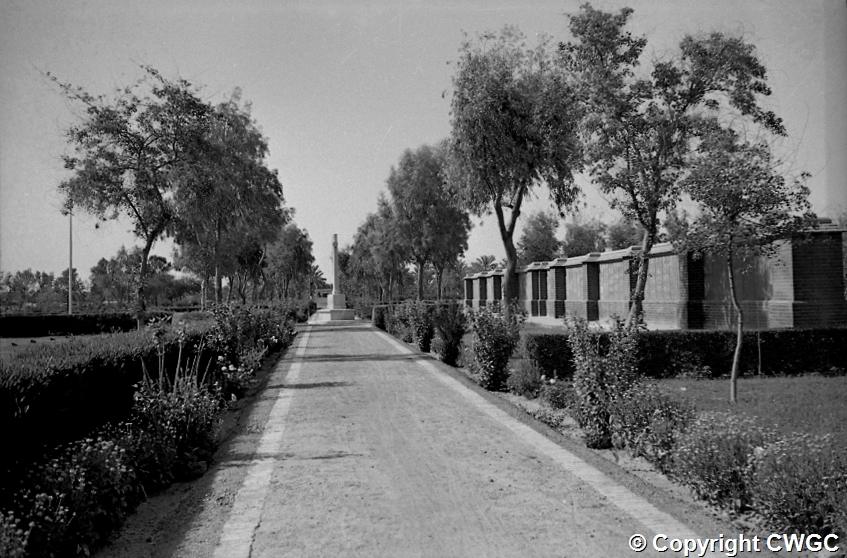 The principal avenue at Basra War Cemetery, looking towards the Cross of Sacrifice in the distance, with the Memorial Wall panels lining the right-hand side and flowering borders edging the path. © CWGC.