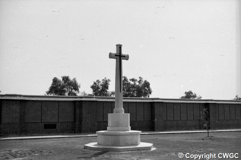 The Cross of Sacrifice on its octagonal pedestal, set against the long sweep of the Memorial Wall and its inscribed panels, with trees rising behind in the open grounds of the cemetery. © CWGC.
