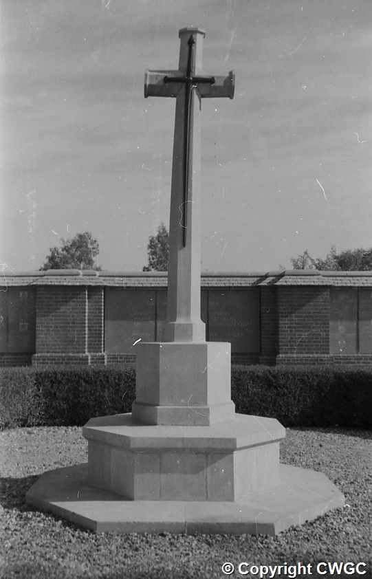A close view of the Cross of Sacrifice on its tiered stone pedestal, with the Memorial Wall panels visible behind hedging and the cemetery trees rising beyond. © CWGC.