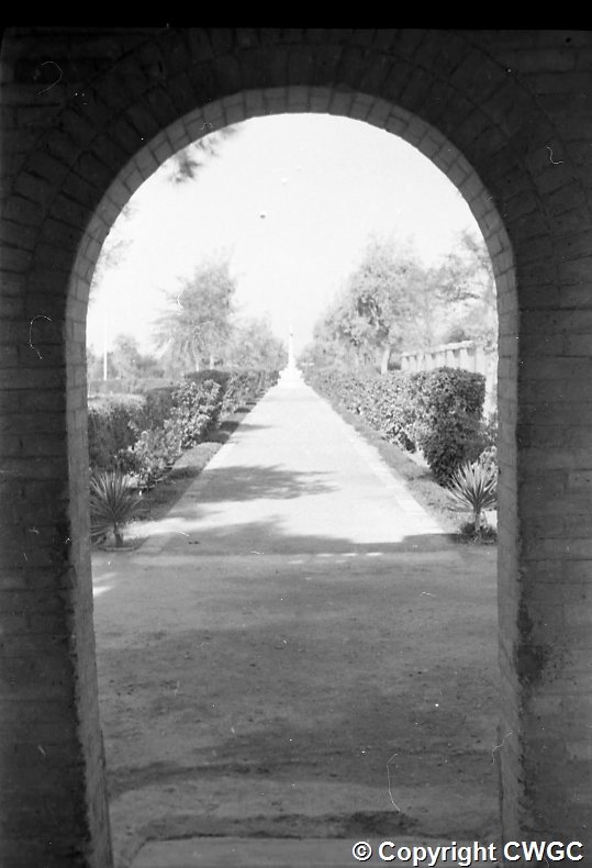 The view through the arched gatehouse entrance, looking down the central path between hedges and planted borders towards the Cross of Sacrifice in the distance. © CWGC.