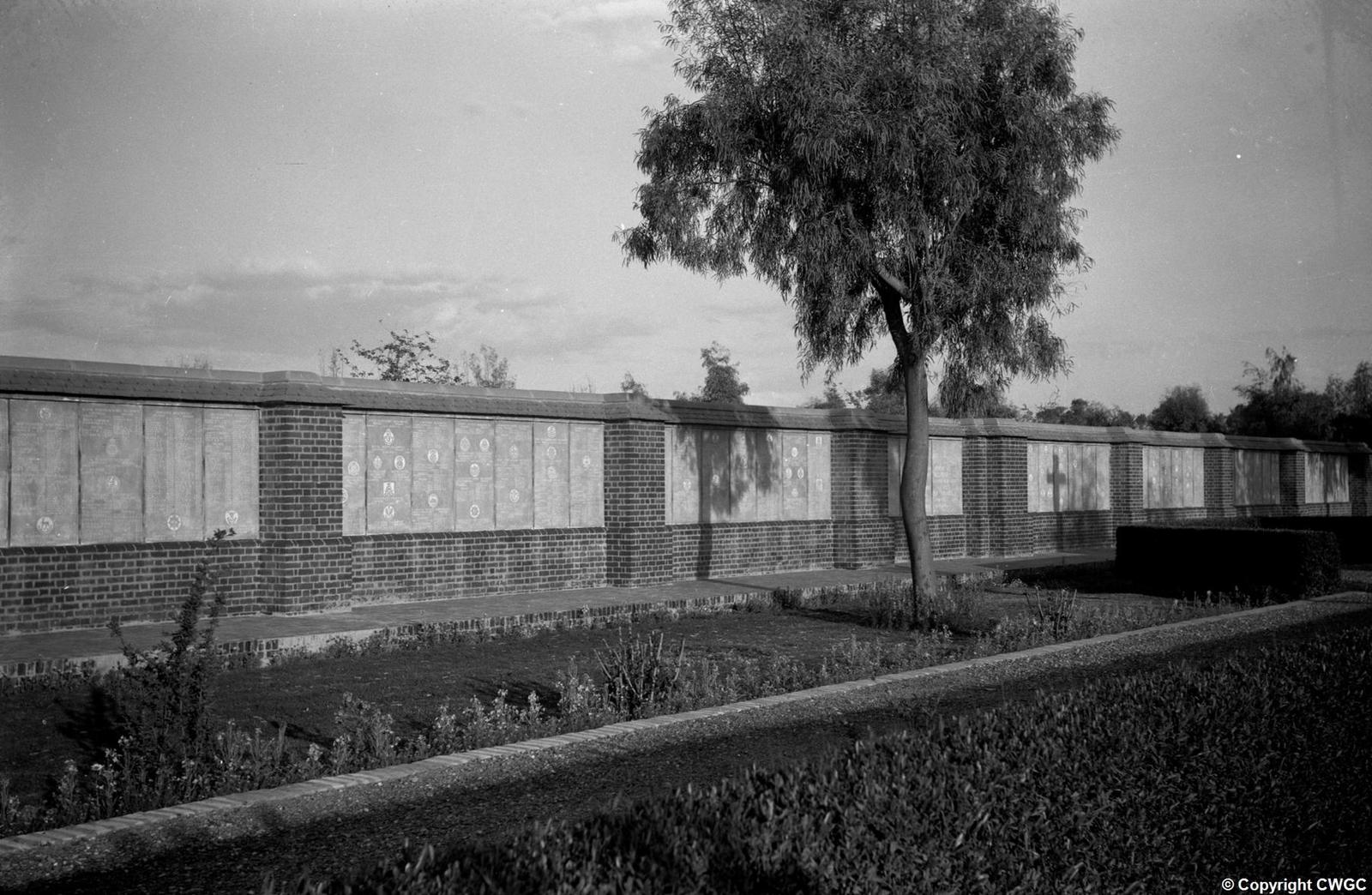 A long run of Memorial Wall panels with their regimental badges and inscribed names clearly visible, set above brick plinths and edged by lawns, hedges and a flowering border. © CWGC.