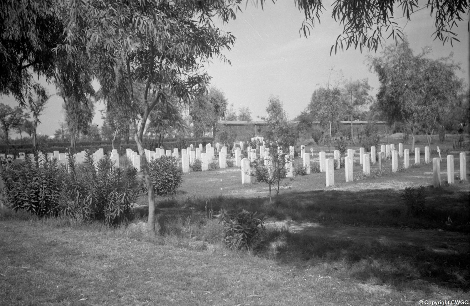 Plot 8 seen from beneath the trees, with rows of headstones leading the eye towards a section of the Memorial Wall in the background, the grounds laid out as a quiet garden. © CWGC.