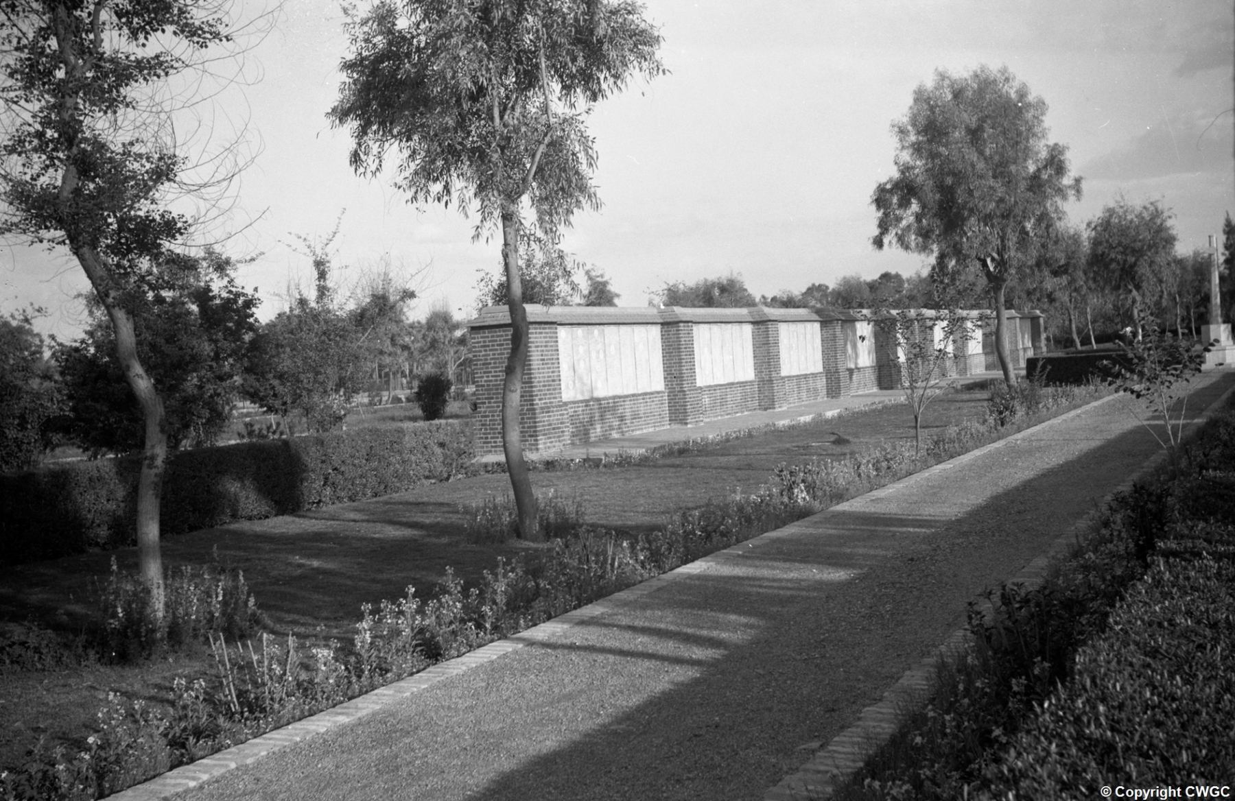 A section of the Memorial Wall set within the cemetery garden, the inscribed panels still bright and intact, with mown lawns, clipped hedges and the Cross of Sacrifice just visible at far right. © CWGC.