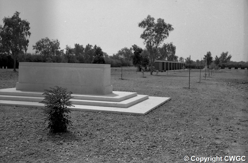 The Stone of Remembrance at Basra War Cemetery in the 1950s, standing on its three tiered platform within the formal cemetery garden, with rows of young trees, a freshly planted shrub in the foreground and one of the brick gatehouses visible in the distance.
