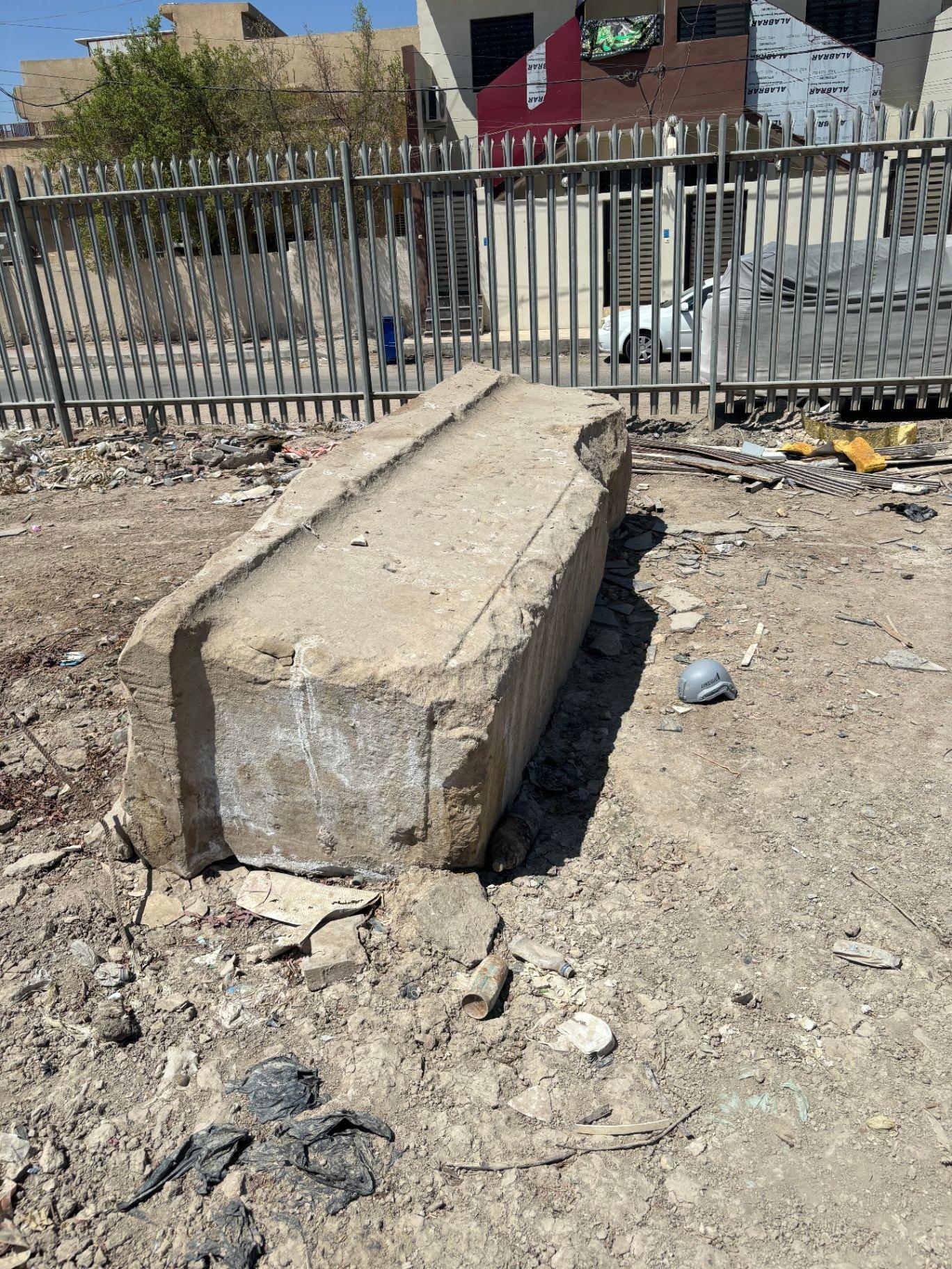 The Stone of Remembrance at Basra War Cemetery today, photographed on the ground beside the perimeter railings with modern Basra visible beyond.