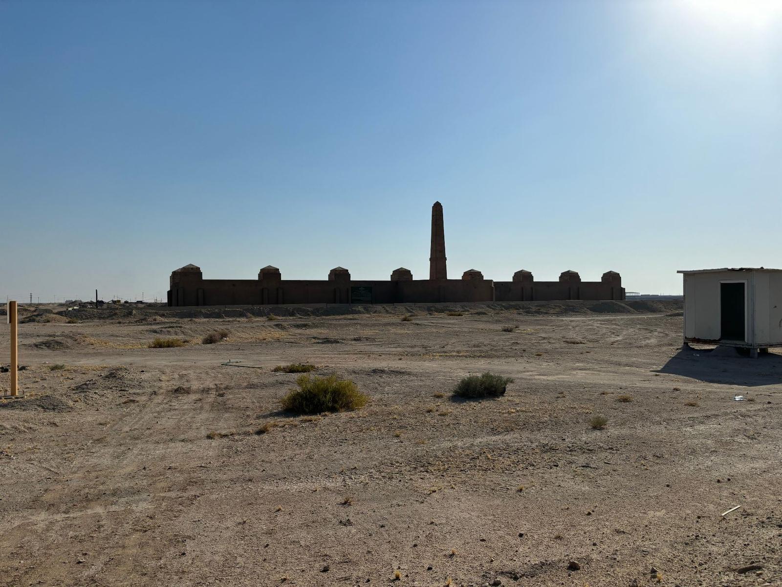 The Basra Memorial seen in silhouette from across the open desert at its relocated site, with the obelisk and pavilions backed against the bright sky and a small guard hut at right.