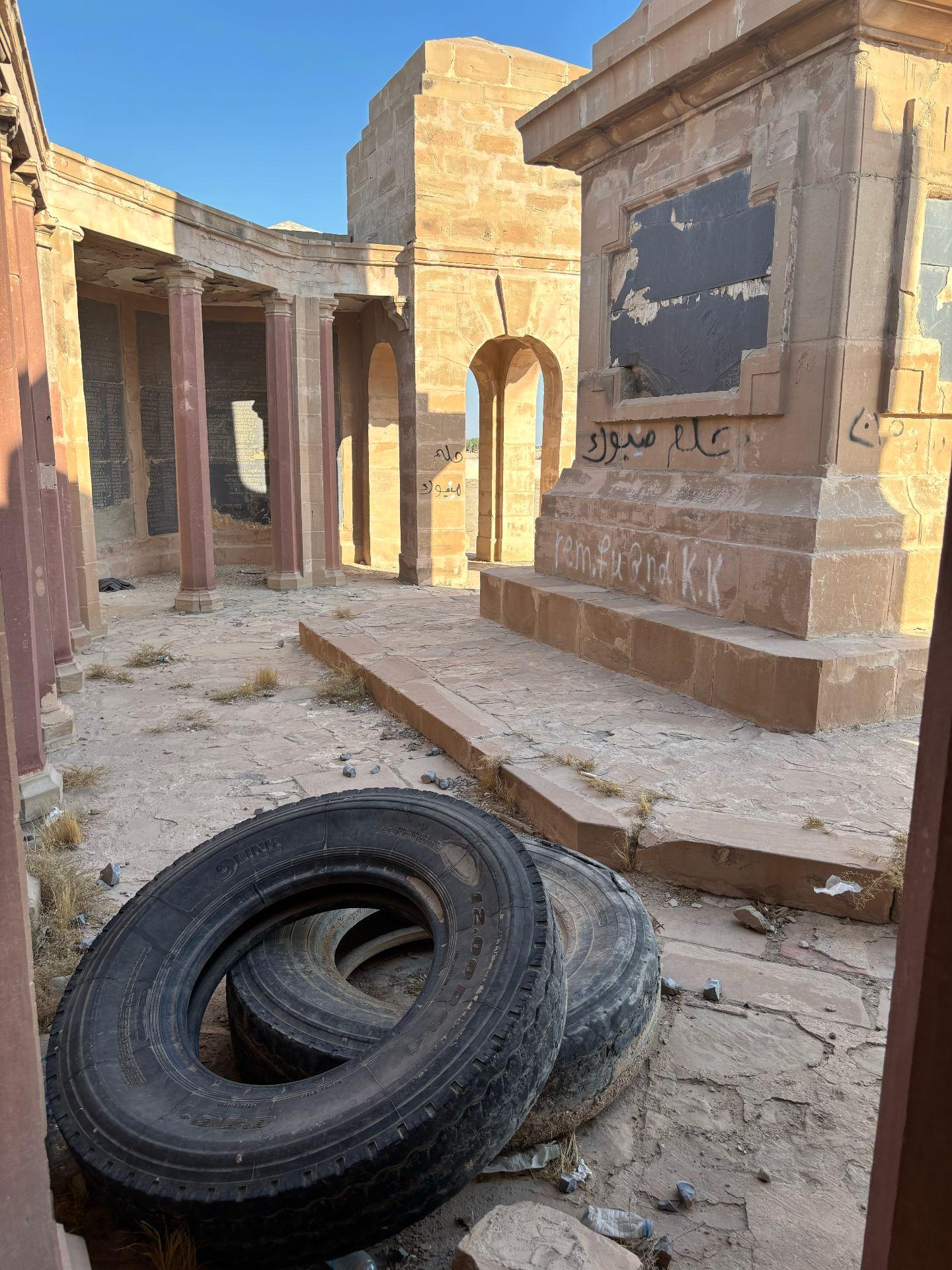 Inside the Basra Memorial today, looking through the colonnade towards the inscribed panels, with Arabic graffiti scrawled across the sandstone, a missing memorial panel leaving a black void in the wall, and discarded lorry tyres dumped on the cracked stone paving.