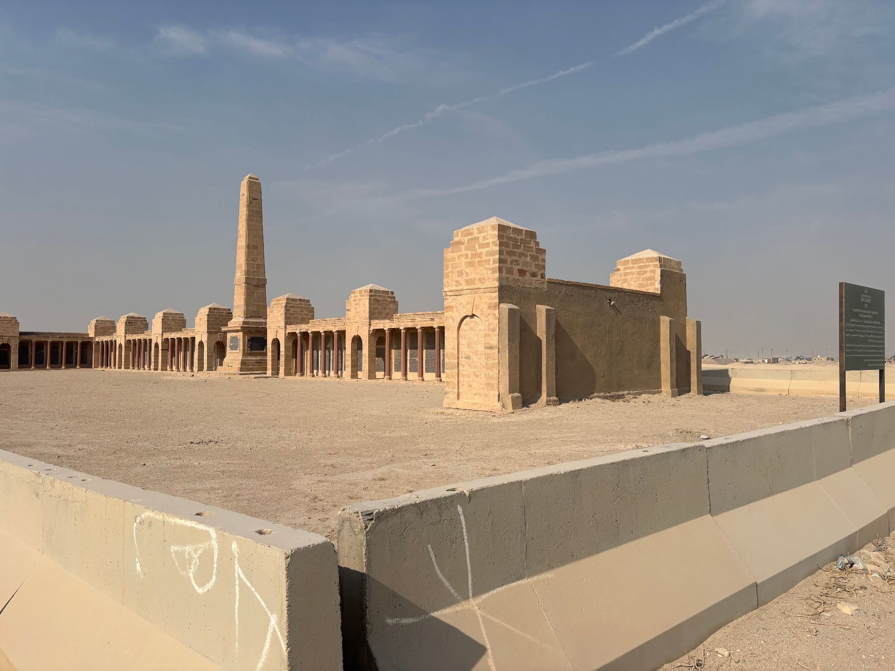 A second view of the Basra Memorial today, showing the obelisk and the full sweep of the colonnaded screen wall on bare desert ground, with the CWGC information panel at right and the perimeter blast wall in the foreground.