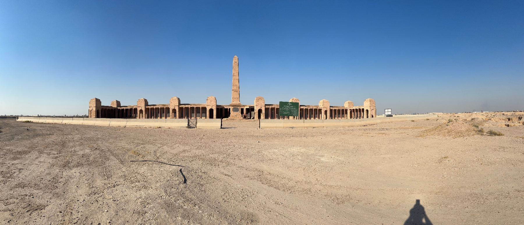 A panoramic view of the Basra Memorial today, the full sweep of the colonnaded screen wall and central obelisk standing alone on the open desert plain.