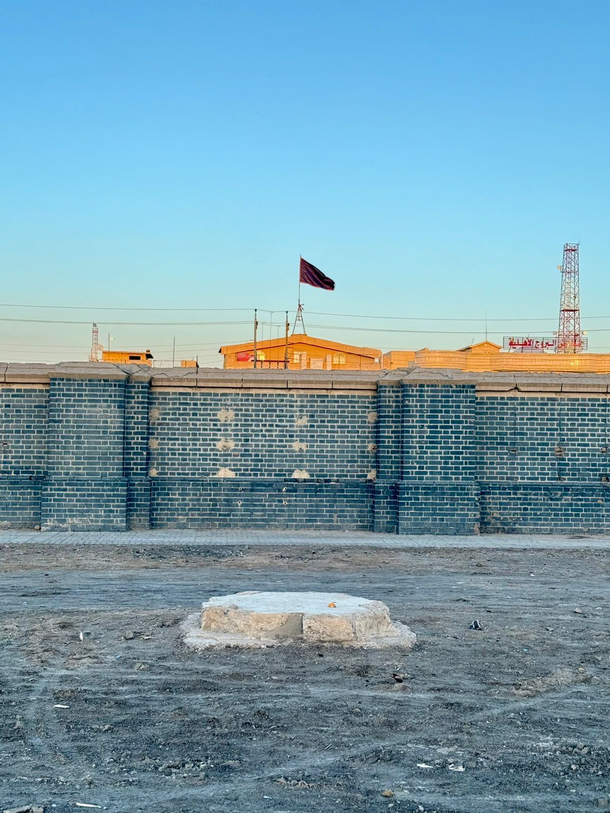 A close view of the stripped Memorial Wall, with the empty recesses where the inscribed panels once stood, an Iraqi religious flag flying beyond the boundary and a single fragment of base remaining in the foreground. Photo: Shane Potaka.