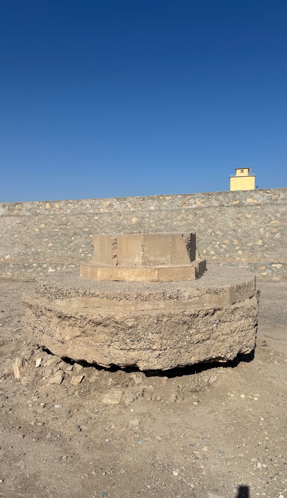 The Cross of Sacrifice pedestal standing on its exposed concrete foundation, the surrounding ground bulldozed away to leave the footing hanging clear of the earth.