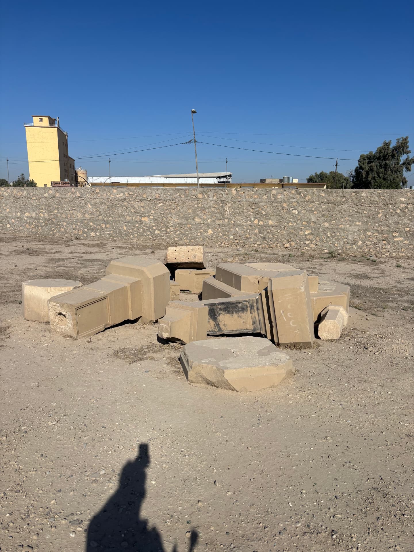 The dismantled memorials lying in a heap of blocks on the cleared ground beside the new perimeter wall, with the grain silo beyond.