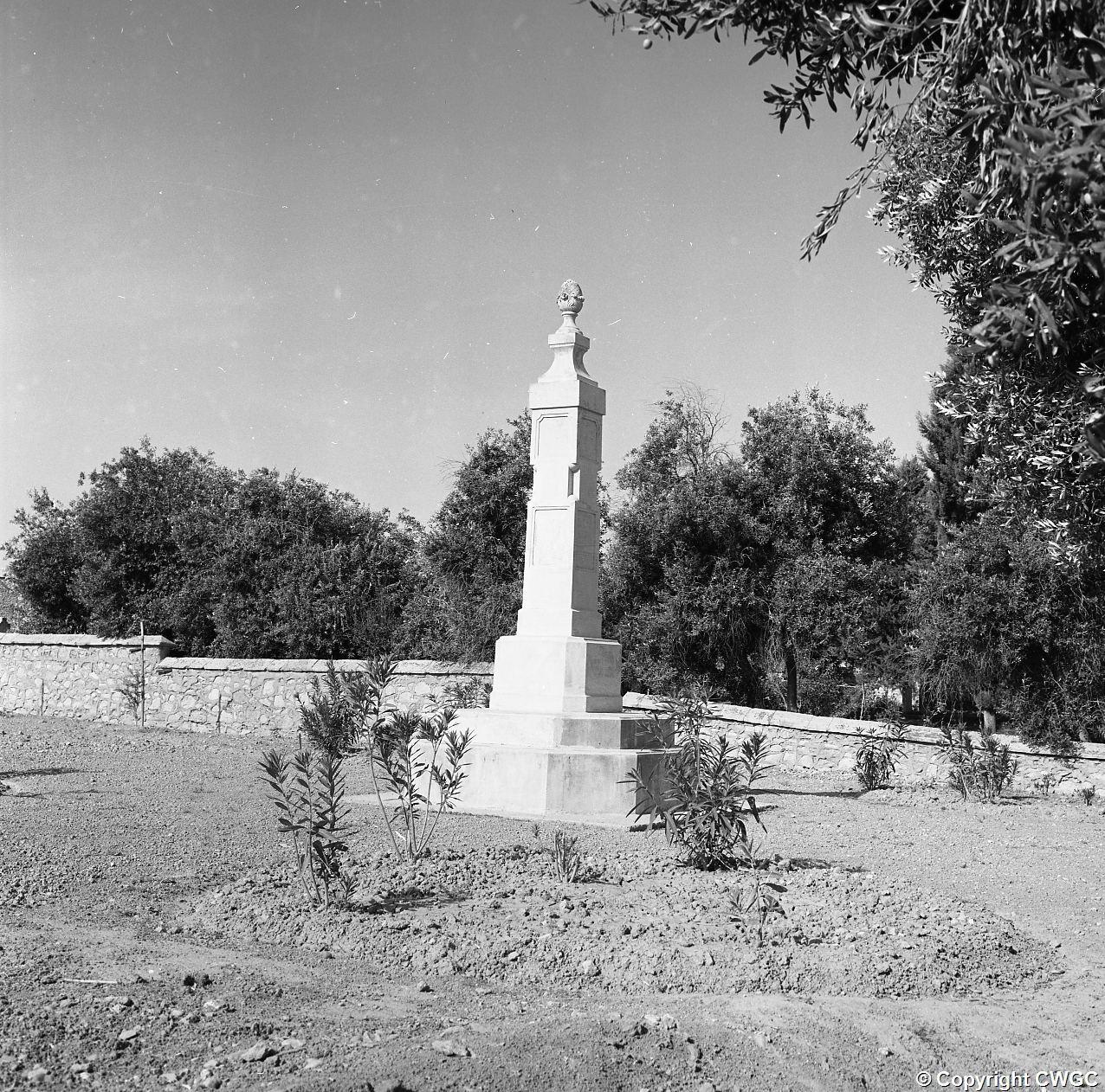 One of the Indian memorial obelisks standing among young planting beside the cemetery wall. © CWGC.