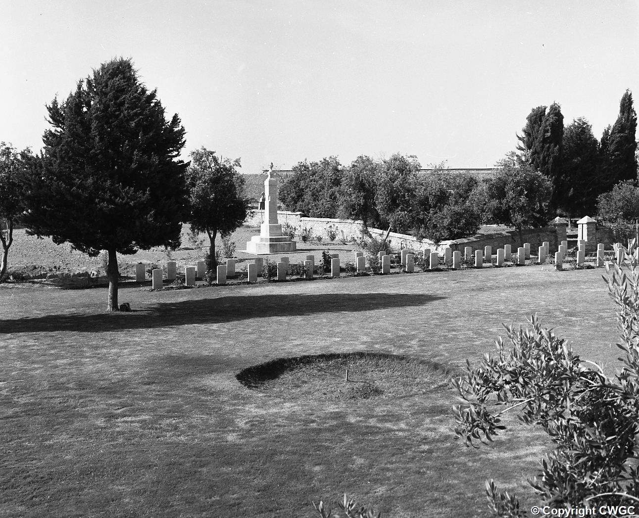 A broad view across the maintained lawn towards the headstones, memorial and mature trees. © CWGC.