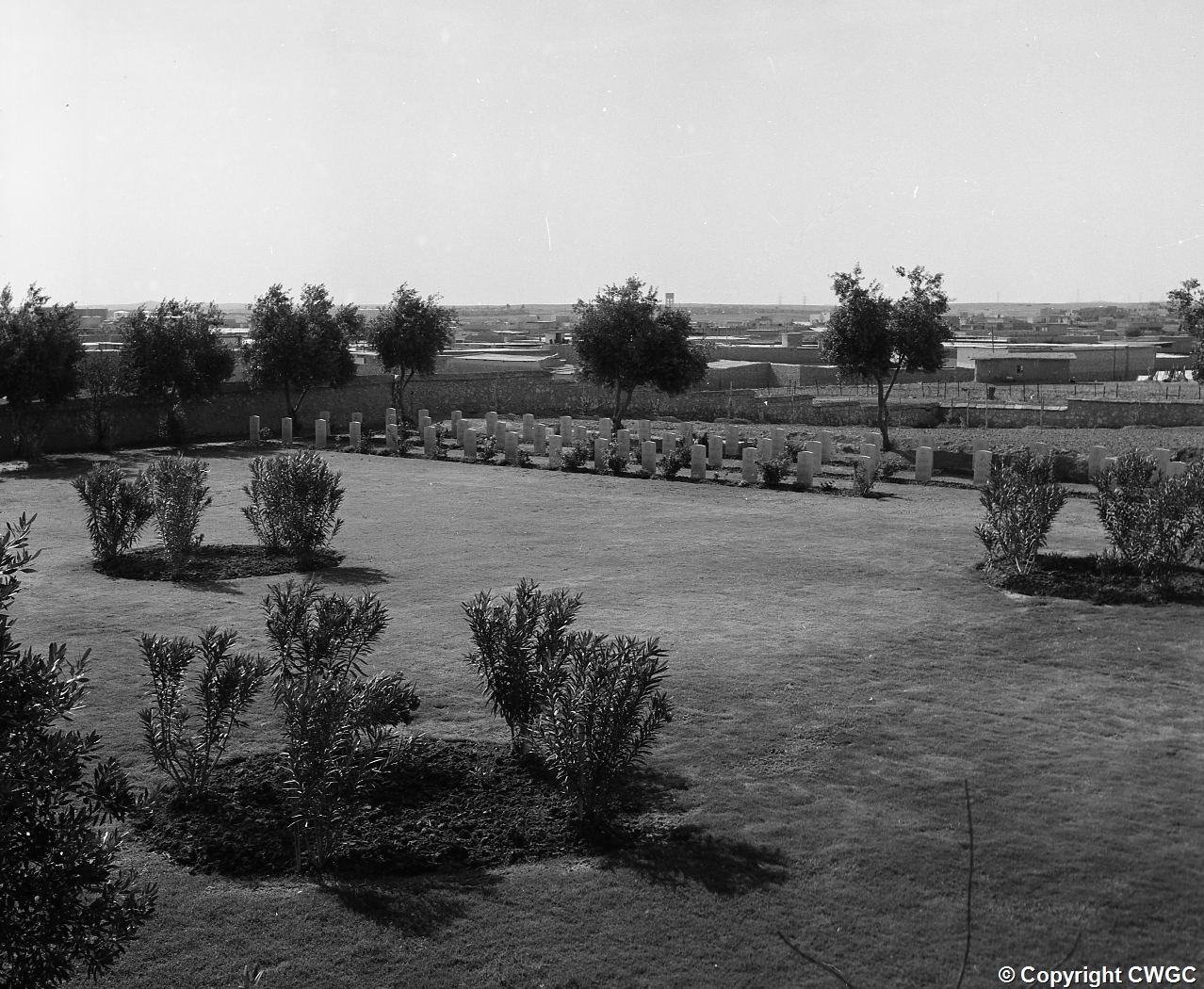 A wider view across the cemetery lawn towards a row of headstones, with the outskirts of Mosul visible beyond the boundary wall. © CWGC.