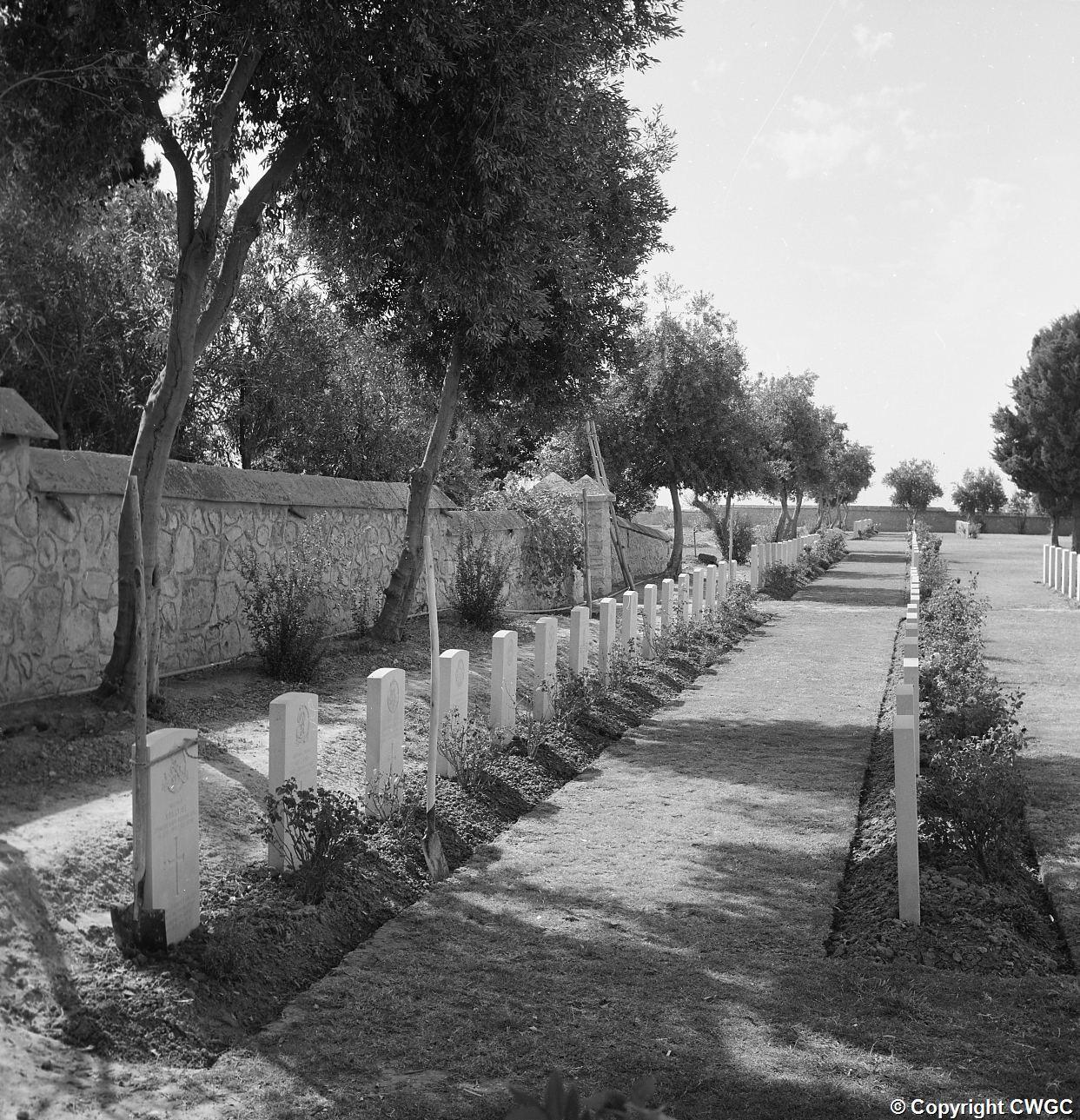 A grass path between planted borders and headstones, shaded by mature trees along the perimeter wall. © CWGC.