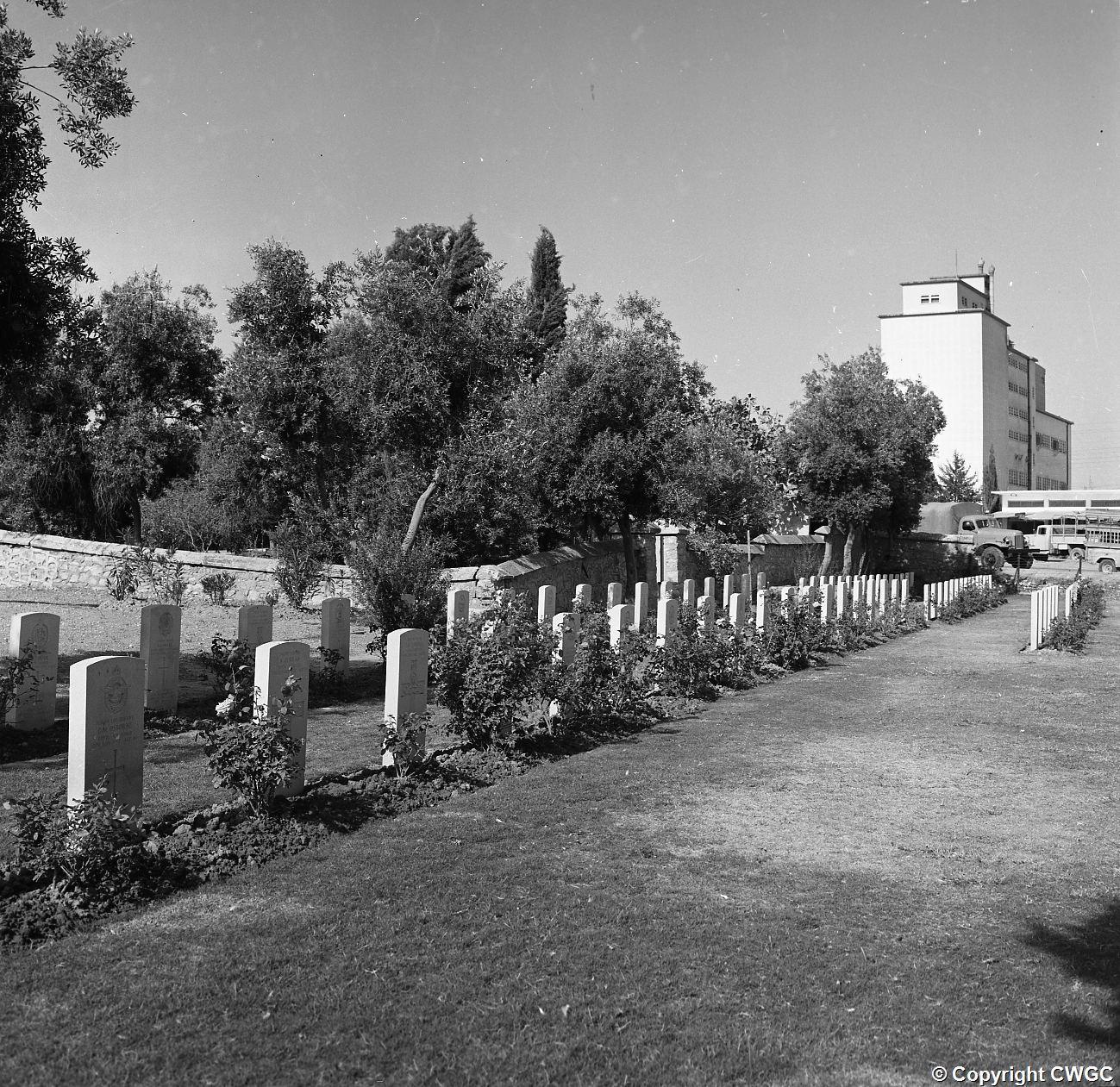 Headstones of RAF and Army casualties in the foreground, with the grain silo and city buildings rising beyond the cemetery wall. © CWGC.