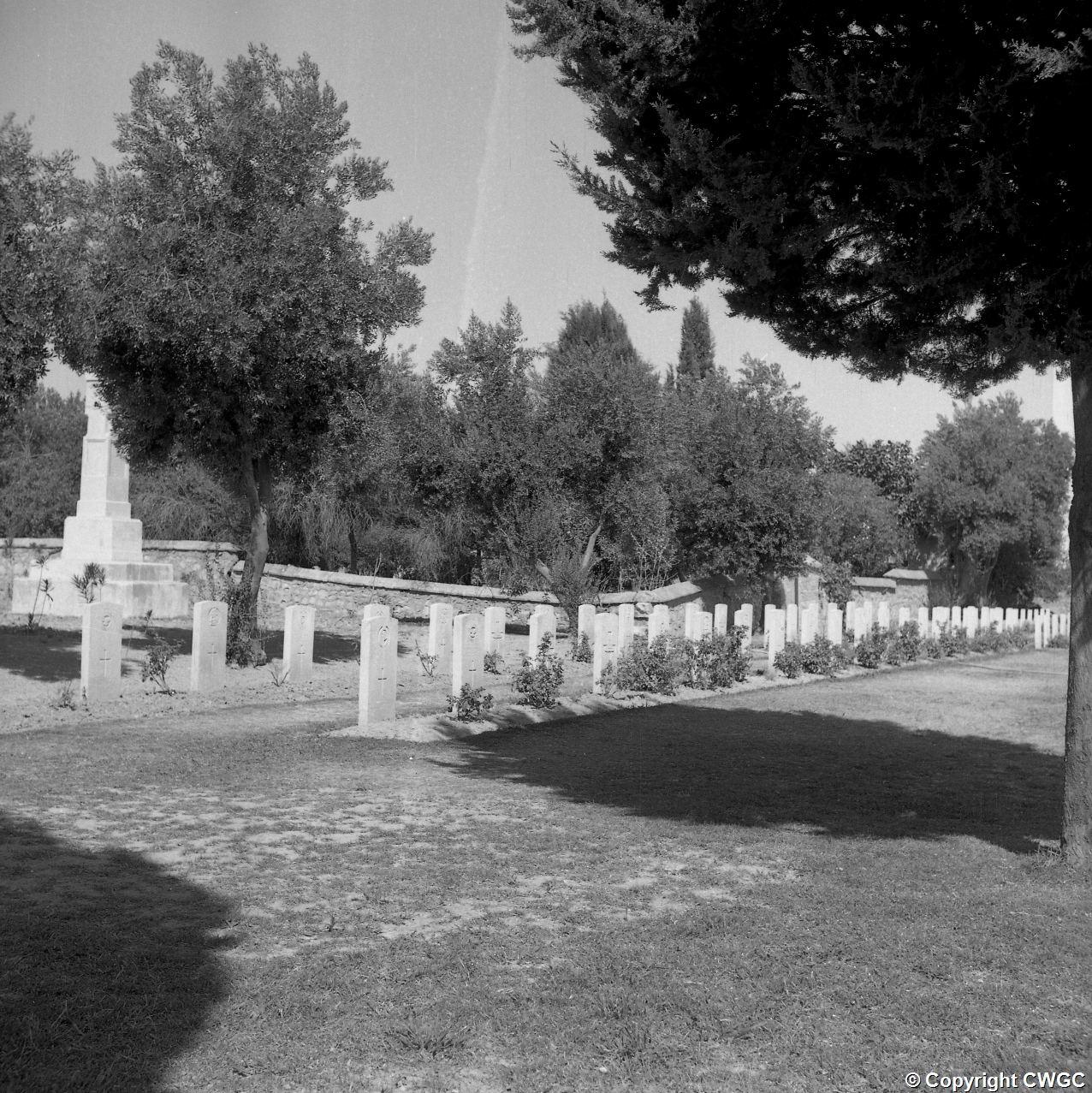 A long row of headstones beneath the trees, with one of the Indian memorial obelisks visible at the left of the plot. © CWGC.
