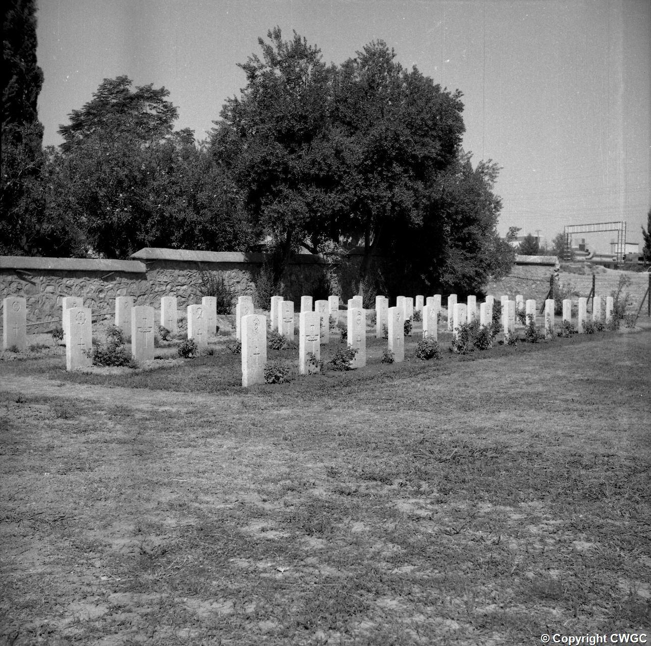 Rows of headstones and planted roses beside the stone perimeter wall. © CWGC.