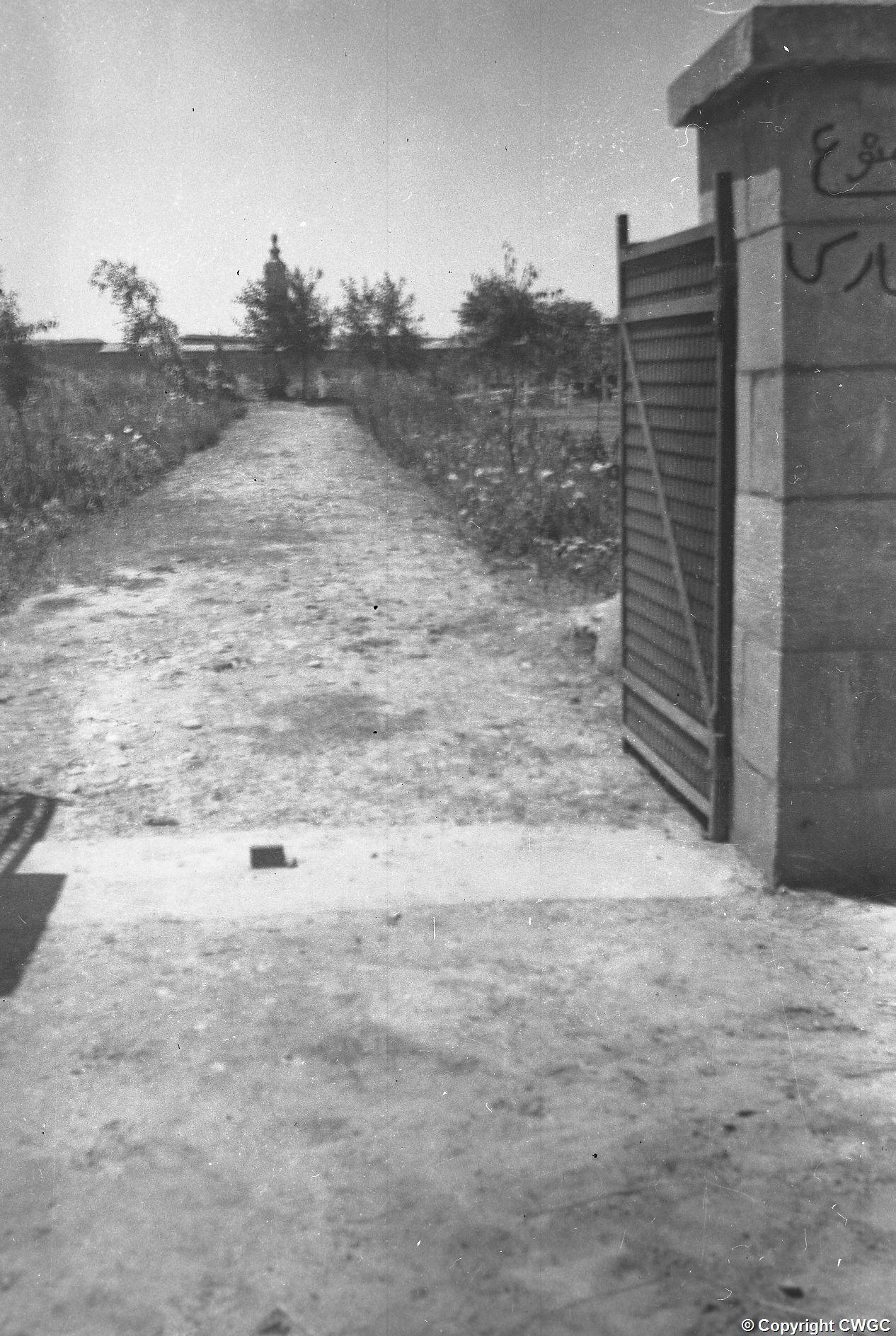 The entrance to Mosul War Cemetery, looking down the path towards the Cross of Sacrifice, with the gate standing open and graffiti on the stone pier. © CWGC.