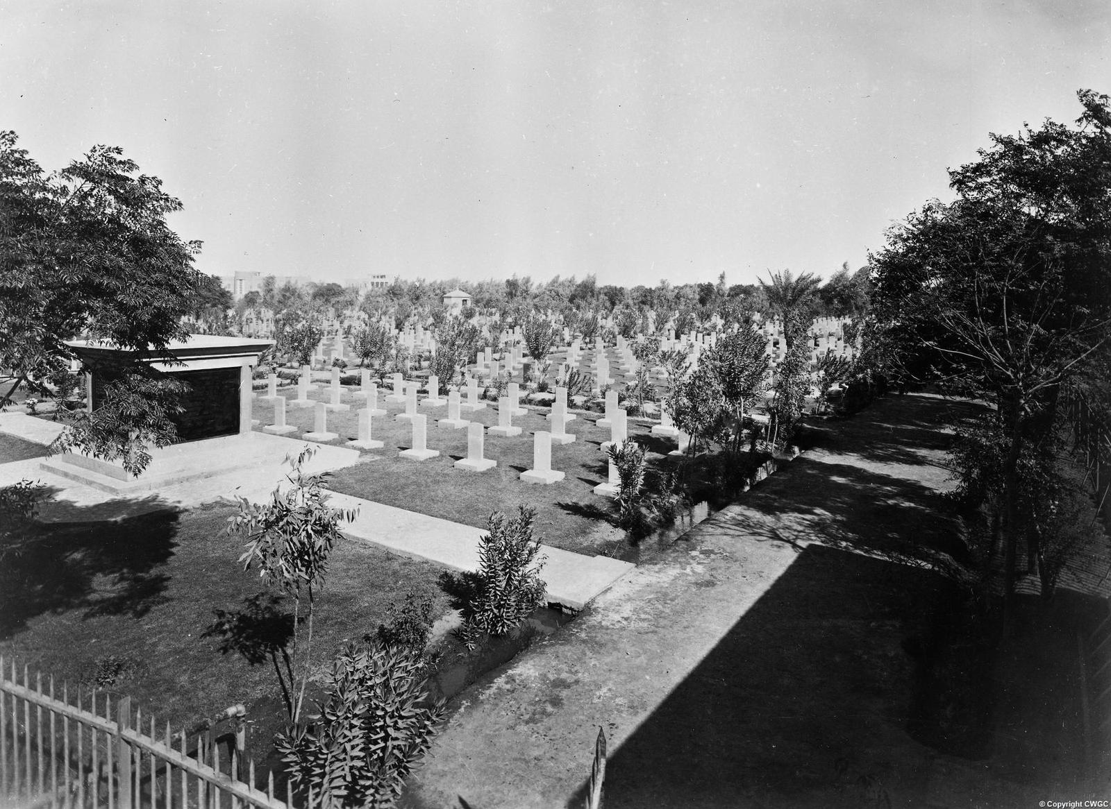 A broad view from the corner of the cemetery, with the 13th Division memorial at left and Maude's tomb in the distance, ordered rows of headstones set in lawns, and the grounds framed by trees and shrubs. © CWGC.
