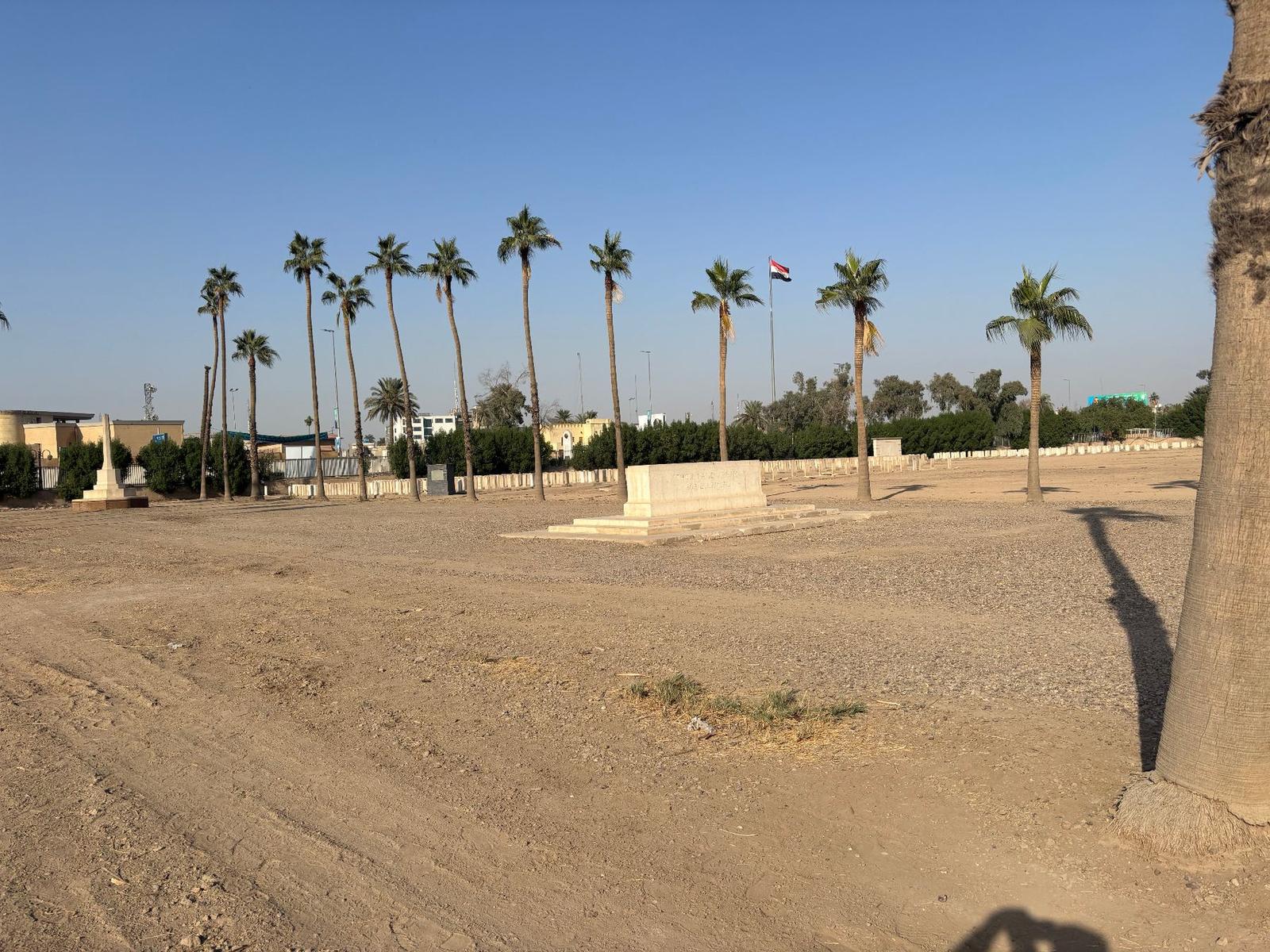 The Remembrance Stone today, set on bare ground beneath a line of date palms, with the Iraqi flag flying beyond the cemetery wall.