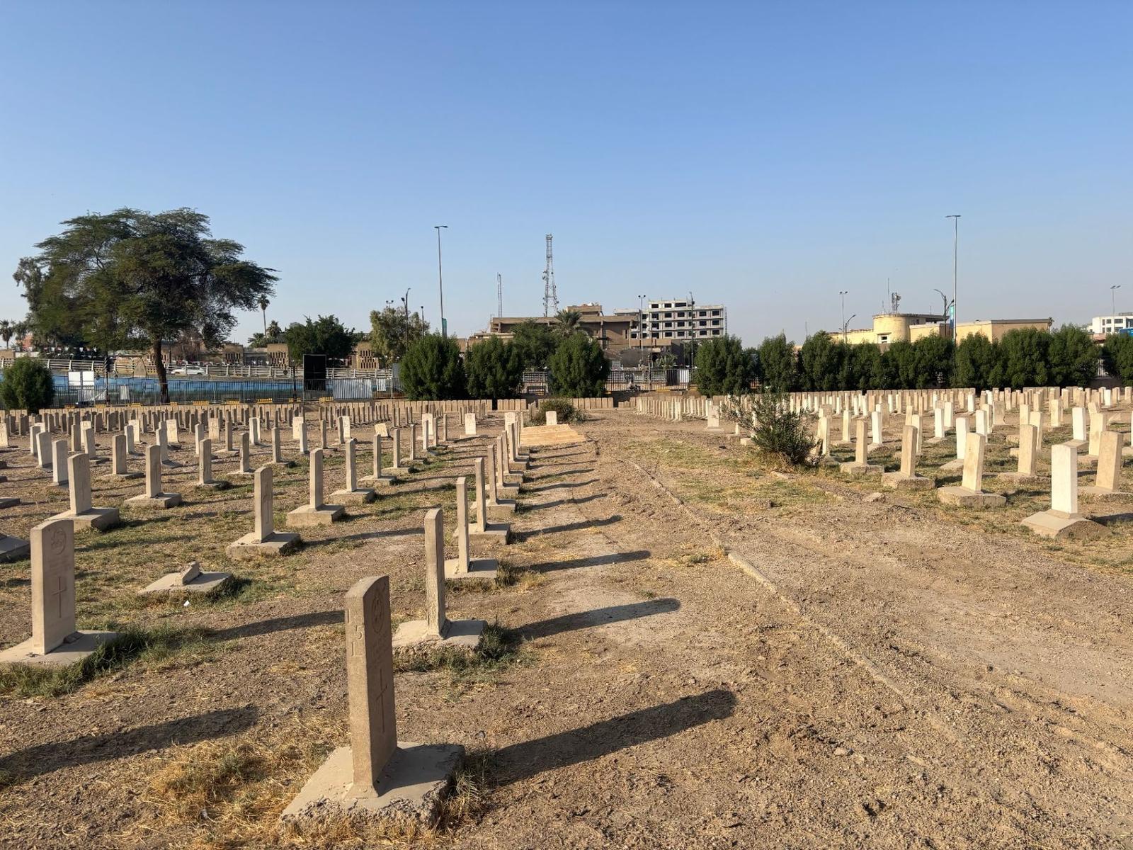 Long rows of headstones receding toward the perimeter wall, with a single tree and the surrounding city visible beyond the cemetery boundary.